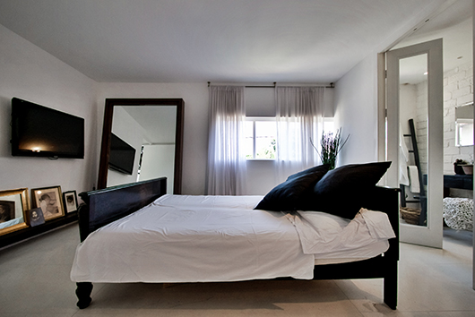 מיטת עץ גדולה במרכז חדר השינה הלבן. מראה גדולה מונחת על הקיר וההשתקפות תורמת לתחושת המרחב | צילום: איתי סיקולסקי