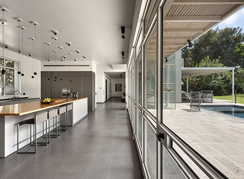 ויטרינות זכוכית ענקיות המחדירות פנימה את נוף החצר האחורית והחורש הירוק שבפאתיה, נמתחות מן הרצפה עד התקרה בכל חללי קומת הקרקע, כולל המטבח וגרם המדרגות | צילום: עודד סמדר